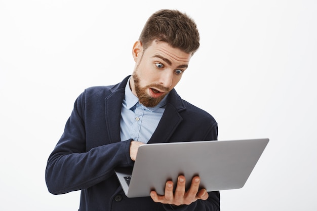 Man going crazy, being in rush working on project. Anxious troubled good-looking male with beard in suit holding laptop looking at computer screen, posing concerned and focused against grey wall