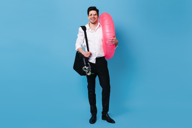 Man in great mood holds inflatable circle. Guy in shirt and pants posing with diving mask on blue space.