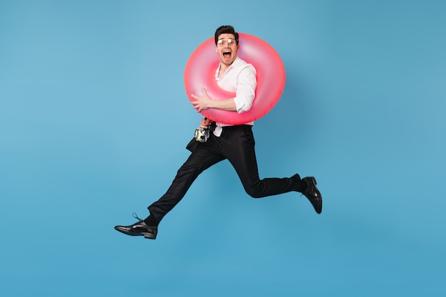 Man in great mood is jumping against blue space with pink rubber ring. Portrait of joyful guy in office outfit.