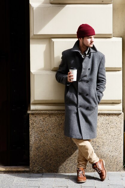 Man in grey coat stands with cup of coffee outside 