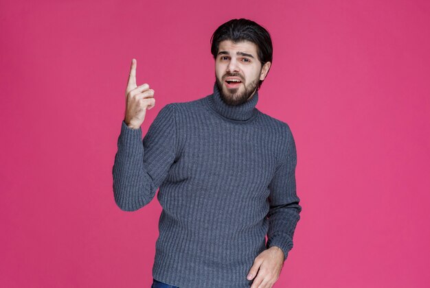 Человек в сером свитере указывает на что-то или представляет кого-то, используя указательный палец.