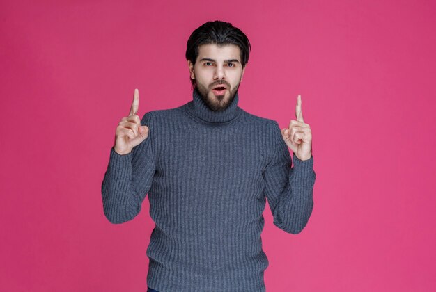 Человек в сером свитере указывает на что-то или представляет кого-то, используя указательный палец.