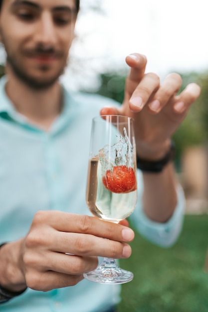 Рука человека бросает клубнику в бокал с игристым вином. Прекрасная жизнь, праздник