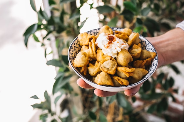 Man hand holds a bowl of fried dushbara dumplings garnished with yogurt and red pepper