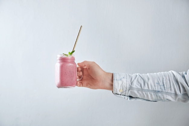 Man hand holds rustic jar with cold fresh berry smoothie with golden straw and mint leves inside. Summer refreshment beverage