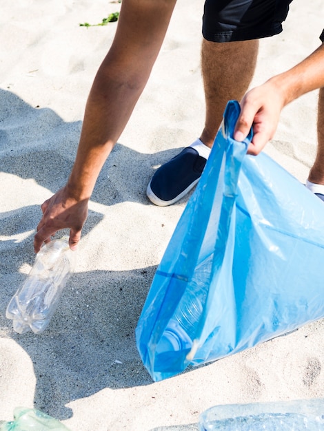 Человек ручной сбор пластиковой бутылки, держа синий мусорный мешок на пляже