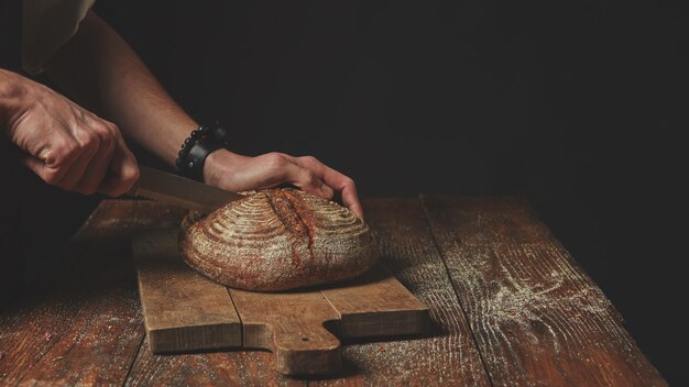 Man hands cut a fresh round bread on a cutting board on a wooden background