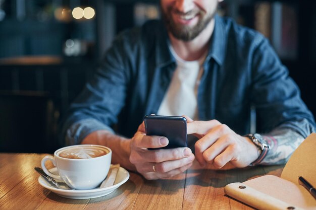 Man hands in denim shirt slide with finger on screen his smart phone, near cup with coffee on wooden table in cafe shop during his coffee break