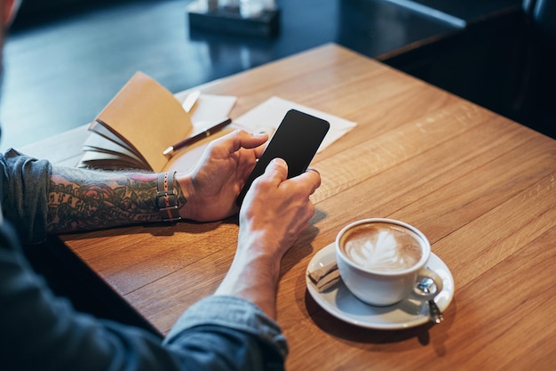 Man hands in denim shirt slide with finger on screen his smart phone, near cup with coffee on wooden table in cafe shop during his coffee break