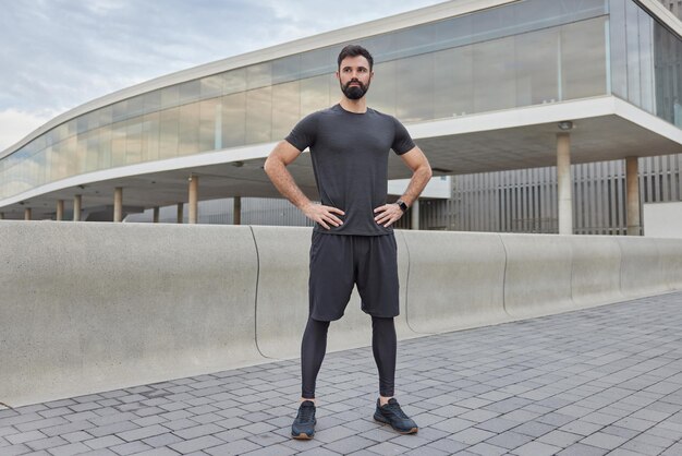 man has regular training outdoors being in good physical shape has muscular arms dressed in sportswear keeps hands on waist maintains healthy lifestyle poses in urban setting