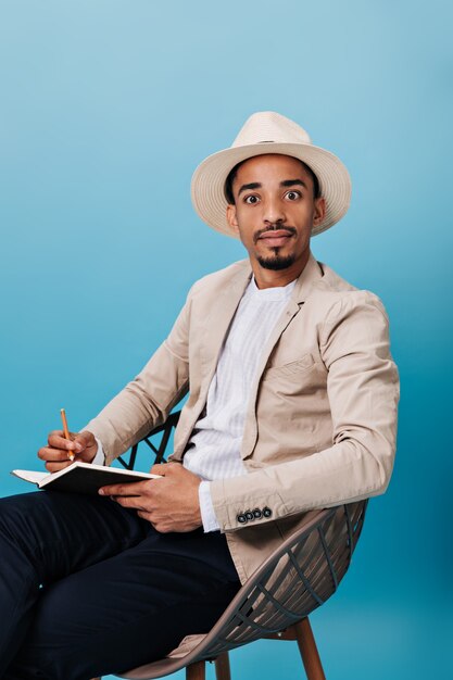 Man in hat and jacket looks at camera in surprise and holds notebook