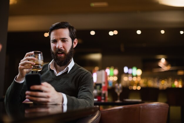 Man having glass of drink while using mobile phone