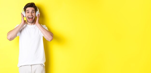 Man in headphones looking surprised and happy listening awesome song standing over yellow background