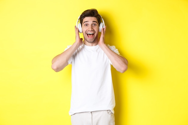 Man in headphones looking surprised and happy, listening awesome song, standing over yellow background.
