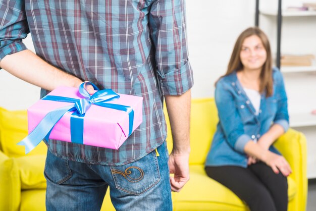 Man hiding gift box behind his back in front of girlfriend sitting on sofa