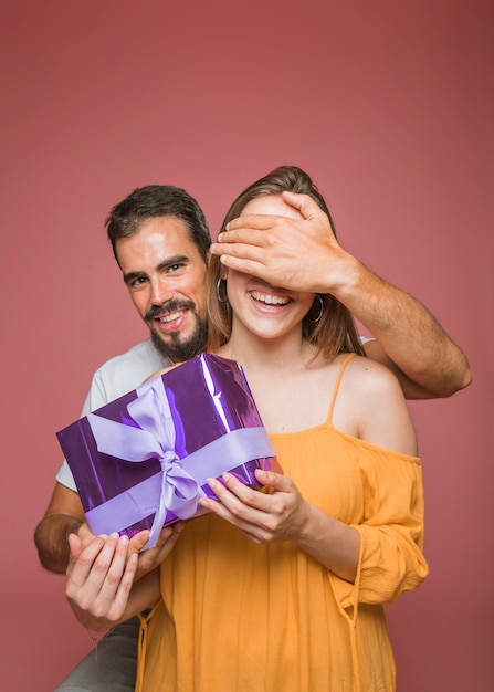 Free photo man hiding her girlfriend's eye holding gift box against colored background