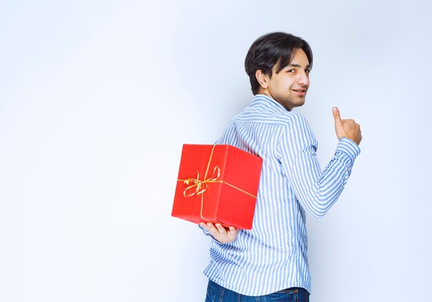 Человек прячет за собой красную подарочную коробку. Фото высокого качества
