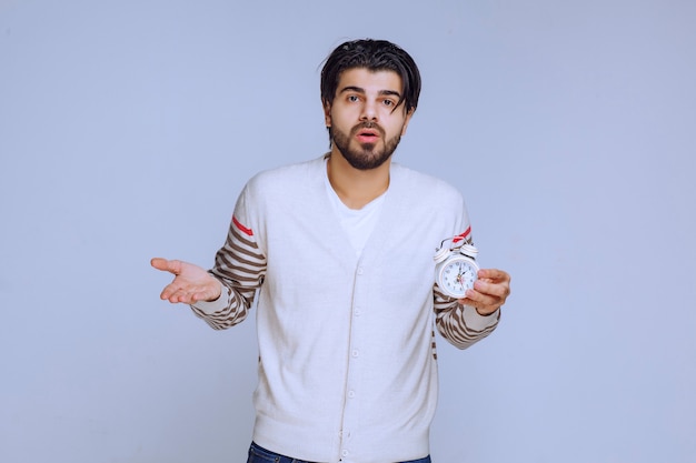 Free photo man holding an alarm clock and looks confused.