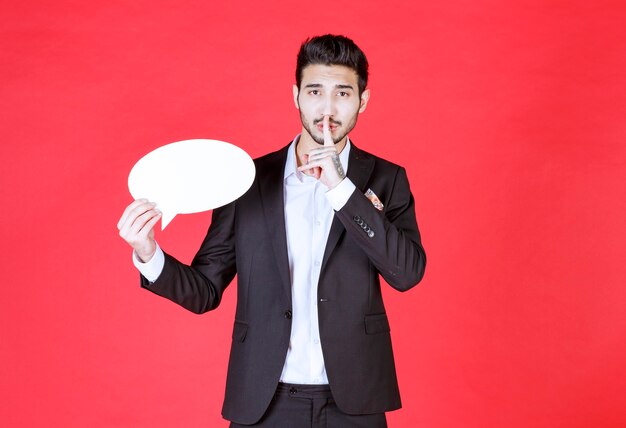 Man holding a blank ovale shape info board and asking for silence.