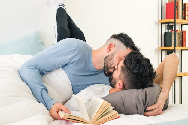 Man holding book in hand kissing his boyfriend lying on bed