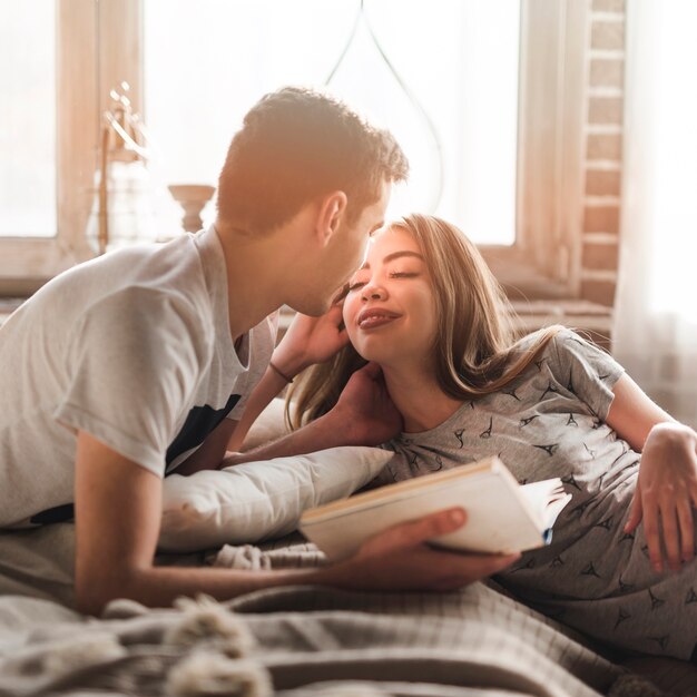 Man holding book in hand looking at woman lying on bed sticking her tongue out