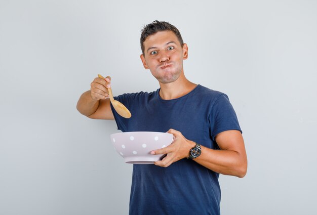 Мужчина держит миску и ложку с надутыми щеками в синей футболке и выглядит голодным. передний план.