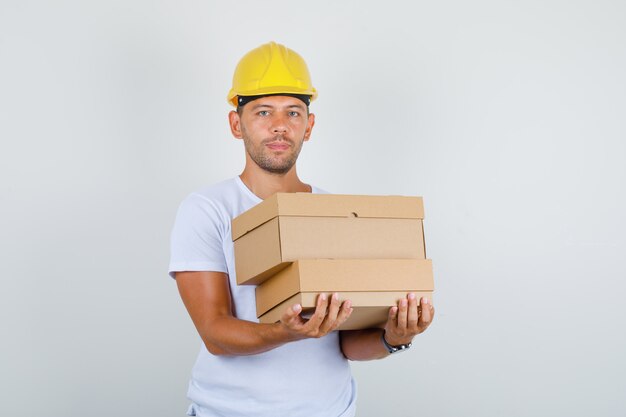 Man holding cardboard boxes in white t-shirt, helmet, front view.