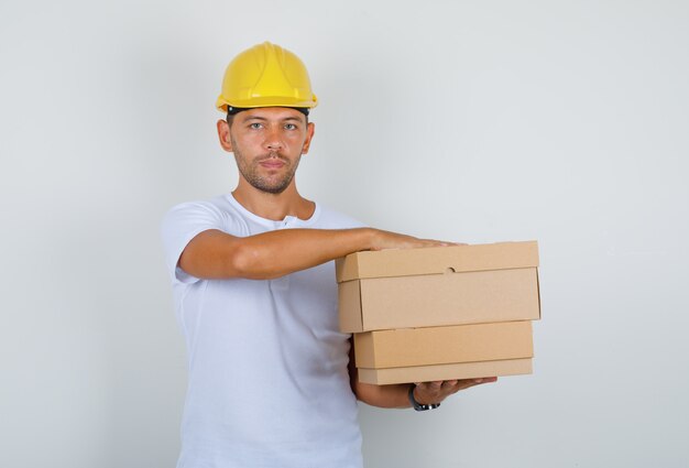 Мужчина держит картонные коробки в белой футболке, шлеме, вид спереди.