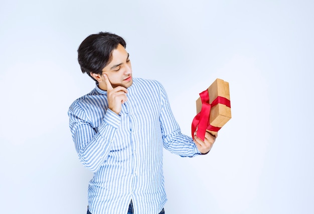 Мужчина держит картонную подарочную коробку с красной лентой и колеблется или смущен. Фото высокого качества