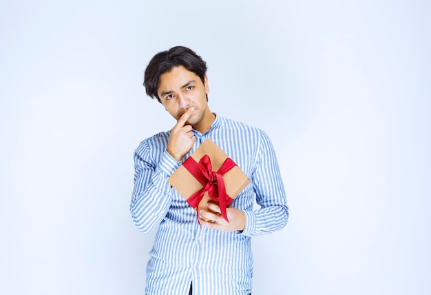 Man holding a cardboard gift box with red ribbon and hesitating or embarassed. High quality photo