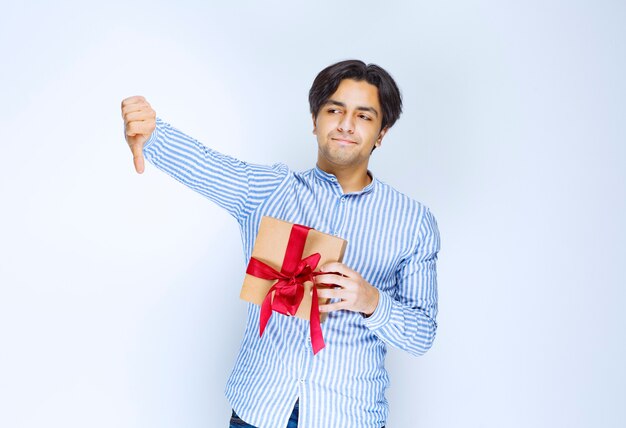 Man holding a cardboard gift box with red ribbon and showing dislike sign. High quality photo