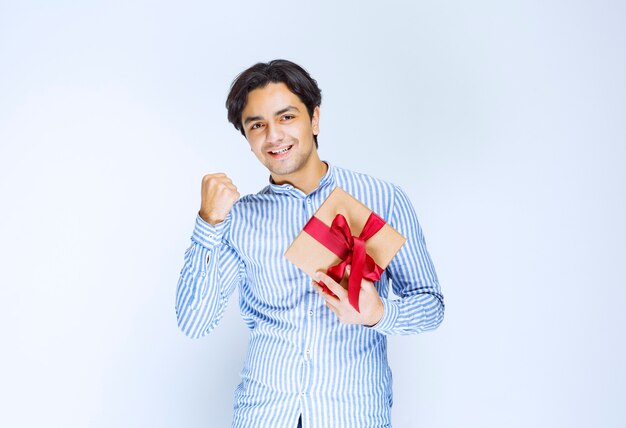 Man holding a cardboard gift box with red ribbon and showing his fist as a successful person. High quality photo