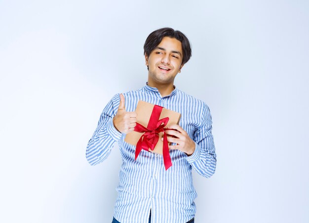 Man holding a cardboard gift box with red ribbon and showing thumb up. High quality photo