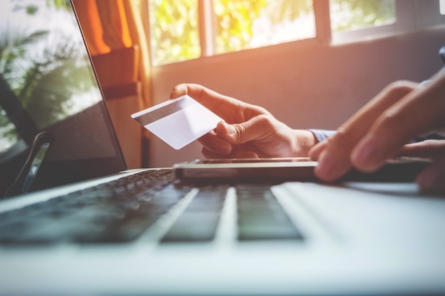 Man holding credit card in hand and entering security code using smart phone on laptop keyboard, online shopping concept.