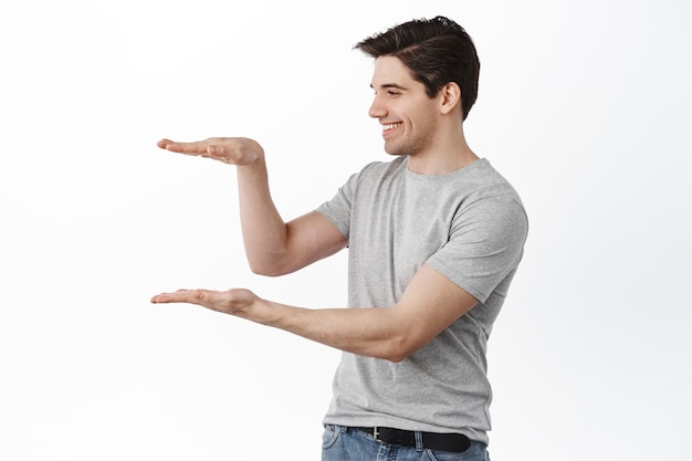 Free photo man holding empty space and smiling, looking at item in his hands with pleased face, standing happy against white wall