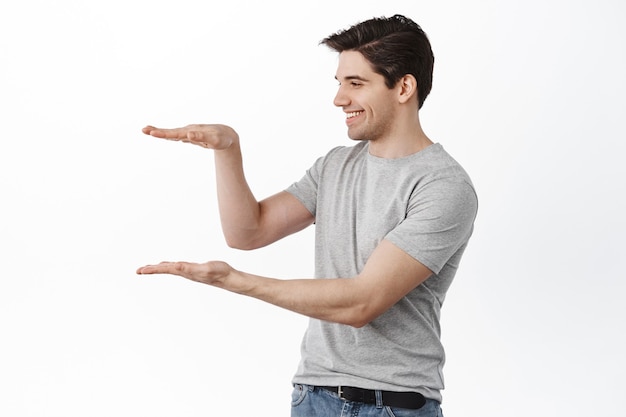 Man holding empty space and smiling, looking at item in his hands with pleased face, standing happy against white wall