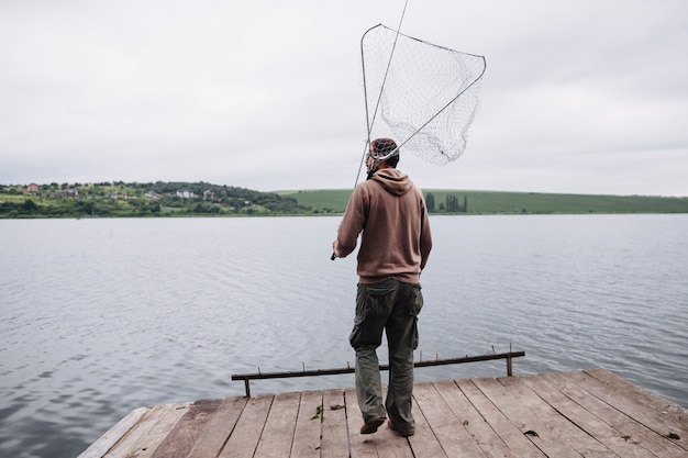 Мужчина, держащий рыболовную сеть и удилище, стоящий на деревянном пирсе перед озером