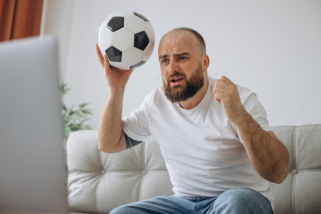 Мужчина держит футбольный мяч и смотрит футбол онлайн дома