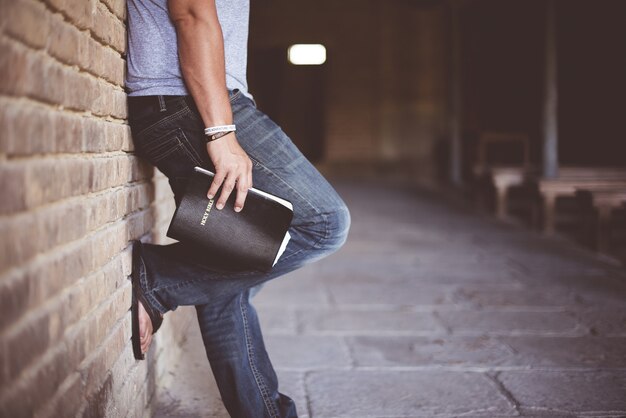 Man holding Holy Bible leaning on bricked wall