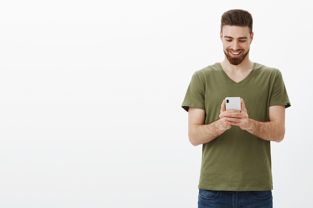 man holding mobile phone looking at device screen with pleased grin, messaging or watching funny amusing video online over white wall