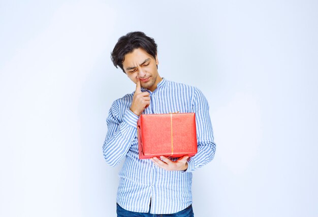 Мужчина держит красную подарочную коробку и выглядит смущенным и задумчивым. Фото высокого качества