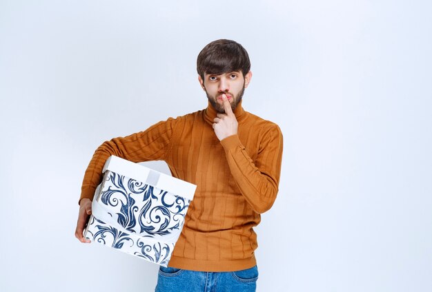 Man holding a white gift box with blue patterns and asking for silence or hesitating about something.