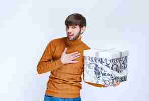 Free photo man holding a white gift box with blue patterns and pointing himself with surprise.