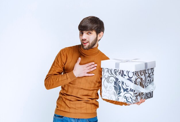 파란색 패턴으로 흰색 선물 상자를 들고 놀라움으로 자신을 가리키는 남자.