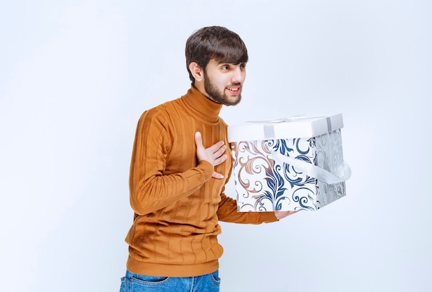 Man holding a white gift box with blue patterns and pointing himself with surprise.