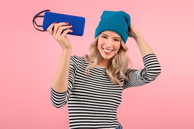 man holding wireless speaker listening to music wearing striped shirt and blue hat smiling posing on pink