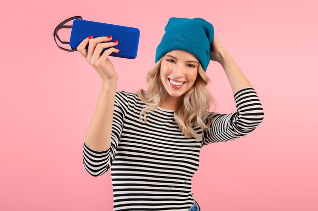 man holding wireless speaker listening to music wearing striped shirt and blue hat smiling posing on pink