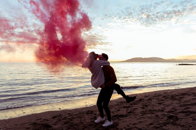 Man holding woman in arms with pink smoke bomb on sea shore