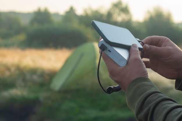 남자는 손에 스마트폰을 들고 자연의 관광 텐트를 배경으로 보조 배터리로 충전합니다. 휴대용 여행용 충전기.