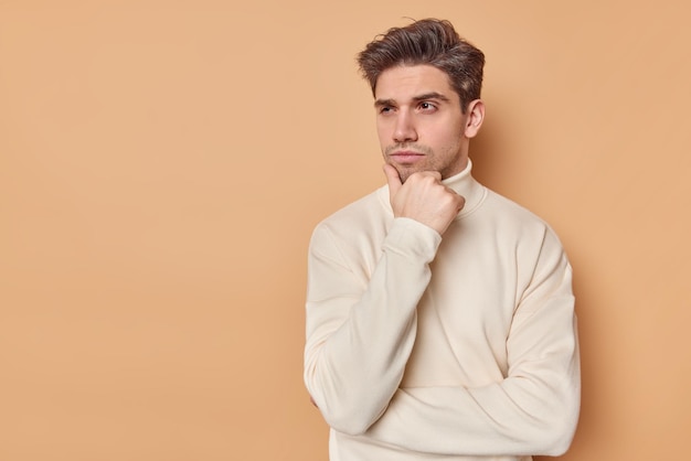 man holds chin looks away pensively concentrated away considers future plans dressed in casual turtleneck isolated on beige with copy space for your text